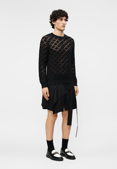 Άνδρας που φοράει μαύρο πουλόβερ με ανοιχτό πλέξιμο, μαύρη φούστα με στρώσεις και λεπτομέρεια δεσίματος, μαύρες κάλτσες και μαύρα-άσπρα μοκασίνια, στέκεται μπροστά σε απλό φόντο.