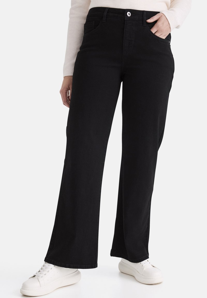 Schwarze Jeans mit weitem Bein aus Denim, mit hohem Bund, Fronttaschen und Knopfverschluss. Kombiniert mit weißen Turnschuhen.