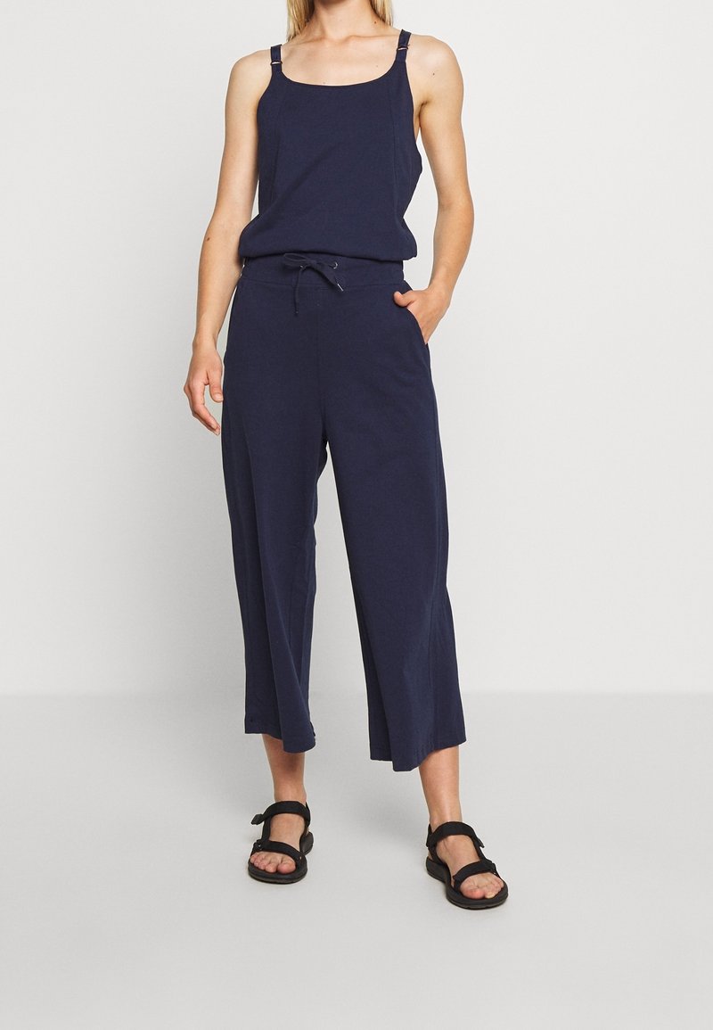 Vrouw draagt een marineblauwe mouwloze jumpsuit met aantrekkoord in de taille en zwarte open-teen sandalen, staand tegen een effen lichte achtergrond.