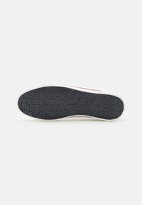 Suela de zapato de goma negra con agarre texturizado. Presenta un acento blanco a lo largo del costado y un patrón de logo en relieve en toda la base.