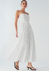 Robe maxi blanche à design à volants, confectionnée dans un tissu texturé avec des motifs ajourés. Bretelles spaghetti et ourlet évasé complètent le look.