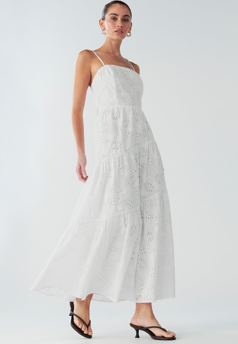 Robe maxi blanche à design à volants, confectionnée dans un tissu texturé avec des motifs ajourés. Bretelles spaghetti et ourlet évasé complètent le look.