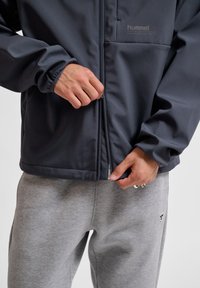 Mörkgrå zip-up-jacka med elastiska muddar, en framficka och slät yta, i kombination med ljusgrå sweatpants med en logotyp.