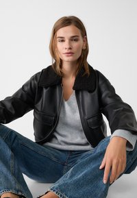 Schwarze Leder-Bomberjacke mit einem strukturierten schwarzen Pelzkragen, ausgestattet mit zwei Fronttaschen, getragen über einem hellgrauen V-Ausschnitt-Pullover. Blaue weit geschnittene Jeans.