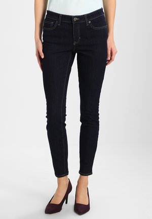 Femme portant un jean skinny bleu foncé et des chaussures à talons hauts violettes, debout devant un fond clair uni.