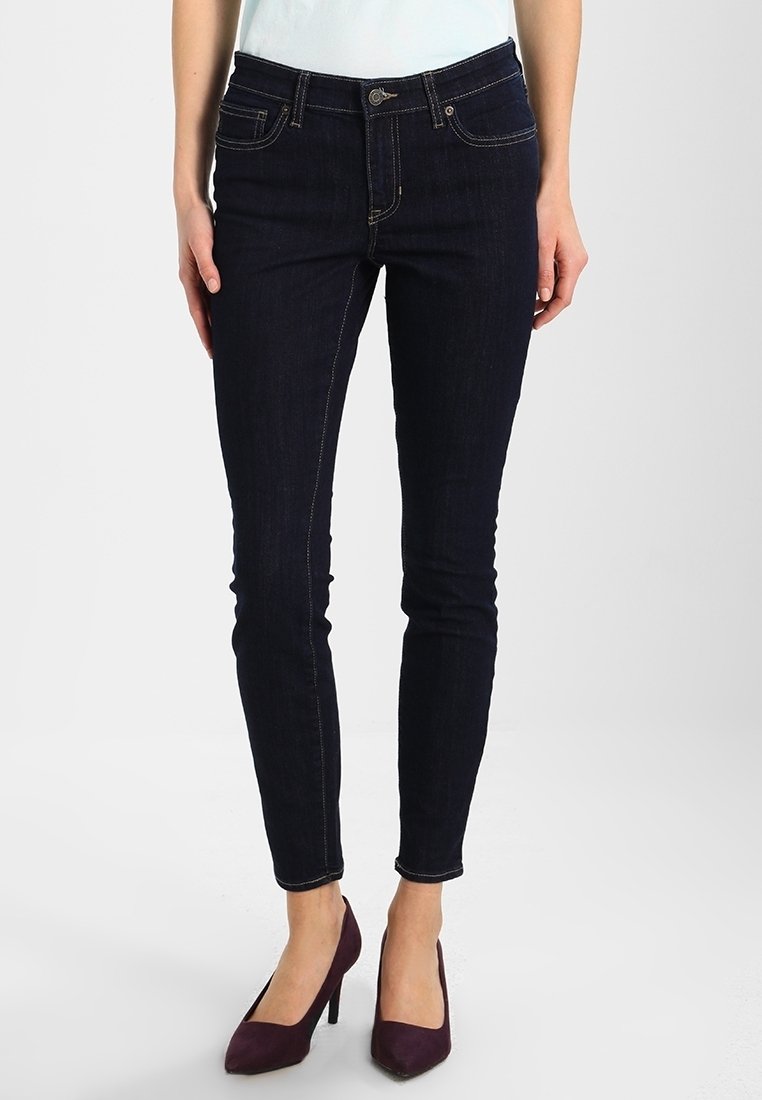 Femme portant un jean skinny bleu foncé et des chaussures à talons hauts violettes, debout devant un fond clair uni.