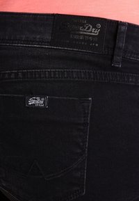 Los jeans de denim negro presentan una cinturilla plana con una parche de marca de cuero, un bolsillo trasero cosido y un diseño minimalista. La vista en primer plano muestra la textura.