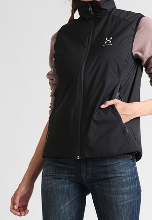 Nainen pukeutunut mustaan hihattomaan vetoketjulliseen liiviin, jossa on logo rinnassa, käsi sivutaskussa, yhdistetty sinisiin farkkuihin ja pitkähihaiseen paitaan.