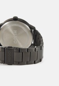 Reloj de pulsera negro con fondo de acero inoxidable, diseño de pulsera enlazada y "HUGO" grabado en el cierre.
