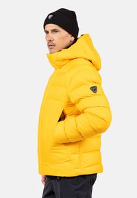 Żółta kurtka puchowa z kapturem, z poziomymi przeszyciami i czarną naszywką z logo na lewym rękawie. Noszona z czarną czapką beanie.