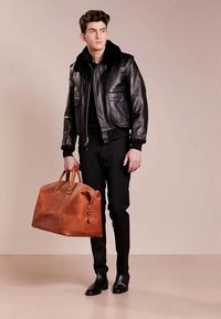 Jauns vīrietis melnā ādas jakā un biksēs, tur holding lielu brūnu ādas sporta somu, stāv uz vienkārša beige fona.