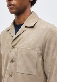 Homme portant une veste en daim beige avec col à crans, trois boutons et une poche carrée sur la poitrine, sur une chemise bleu marine foncé.