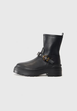 Zwarte leren enkelboot met een chunky zool, voorzien van een zijriem en details in goudkleurige gesp. Gladde textuur en ontwerp met een ronde neus.