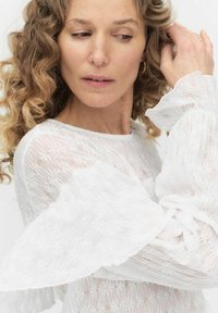 Vit spetsblus med volangärmar i lång modell och blommönster, texturerad tyg och rund halsringning, bärs av en person med lockigt hår.
