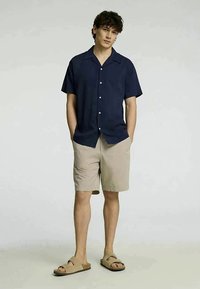 Ναυτικό κοντομάνικο πουκάμισο με κουμπιά, μπεζ σορτς και καφέ παντόφλες. Το πουκάμισο έχει άνετη εφαρμογή και τα σορτς είναι στο γόνατο με λεία υφή.