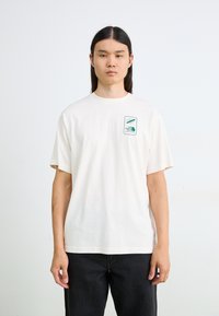 Kortærmet, cremefarvet t-shirt lavet af bomuld. Har et grønt logo-patch på venstre brystområde. Løs pasform med en rund halsudskæring.