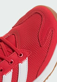 Baskets de sport rouges avec une tige en mesh, des accents en suède et des rayures blanches. Surface texturée avec une semelle en caoutchouc et des œillets renforcés.