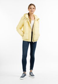 Gul polstret jakke med hætte, frontknapper og sidelommer, parret med tætsiddende jeans og blå sneakers. Glat tekstur, afslappet design.