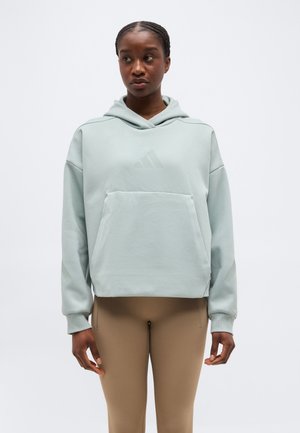 Γυναίκα που φοράει ανοιχτό μπλε φούτερ με τσέπη μπροστά και μπεζ παντελόνι, στέκεται μπροστά σε έναν απλό λευκό φόντο, κοιτώντας προς τα πλάγια.