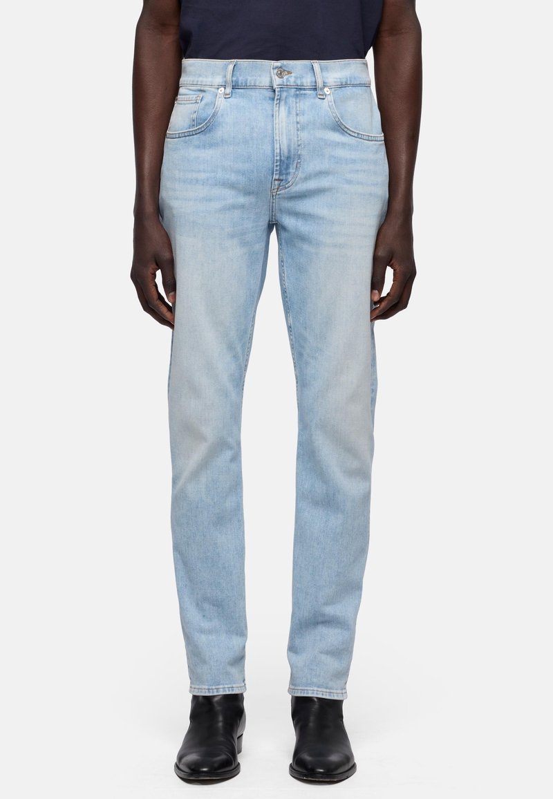 Uomo che indossa jeans di denim azzurro chiaro e stivaletti in pelle nera, in piedi davanti a uno sfondo bianco uniforme.