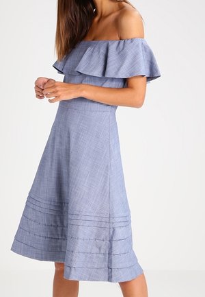Vrouw draagt een lichtblauwe off-shoulder jurk met ruches en een geplooide rok, staand met gevouwen handen voor zich.
