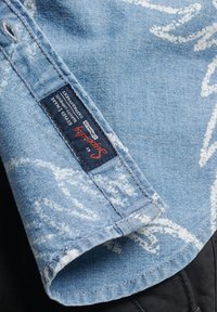 Hellblaues Jeanshemd mit weißen Wellenmustern, einem seitlich aufgenähten Etikett, umgelegtem Kragen und sichtbaren Nähten.