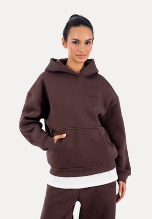 Donna che indossa una felpa con cappuccio marrone scuro e pantaloni abbinati, con le mani nelle tasche della felpa, in piedi davanti a uno sfondo chiaro e uniforme.