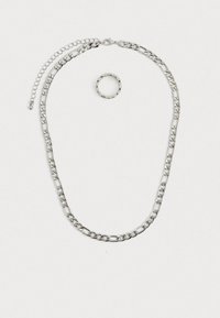 Conjunto de joias em tom prateado, apresentando um colar com corrente grossa e fecho tipo lagosta, além de um anel texturizado que o acompanha. Ambas as peças são brilhantes e metálicas.