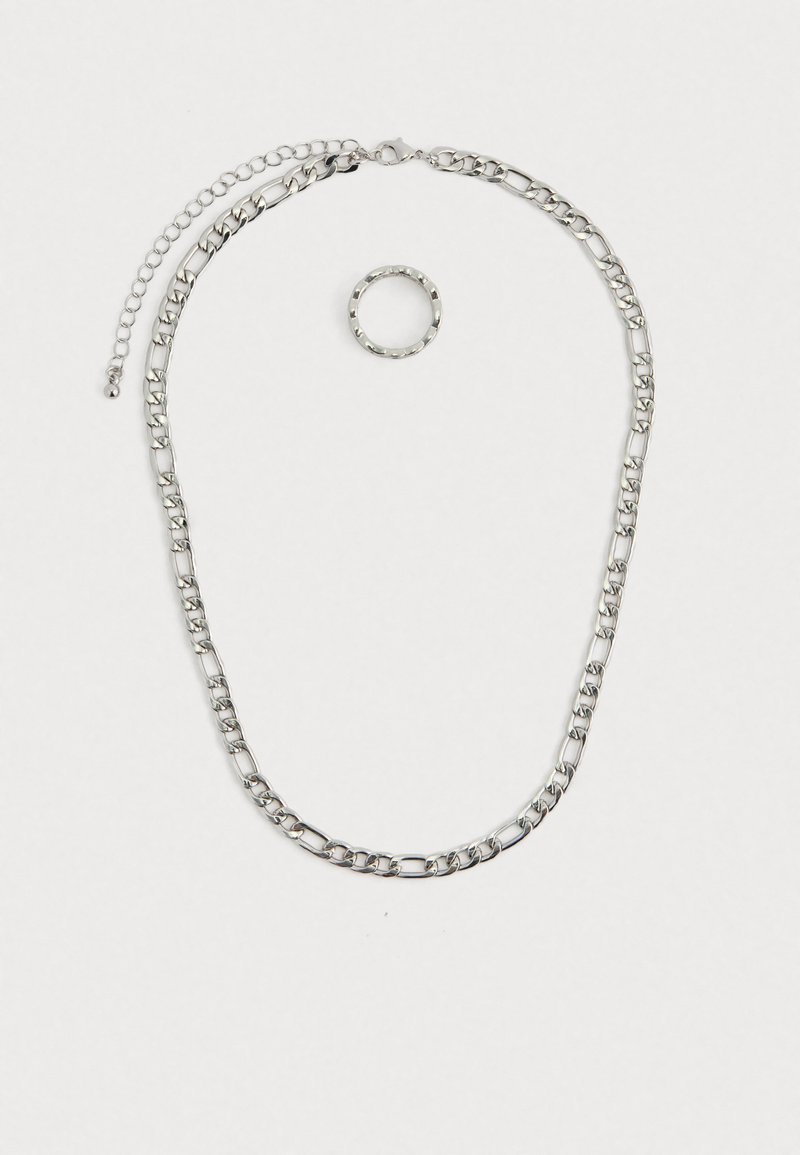 Conjunto de joias em tom prateado, apresentando um colar com corrente grossa e fecho tipo lagosta, além de um anel texturizado que o acompanha. Ambas as peças são brilhantes e metálicas.