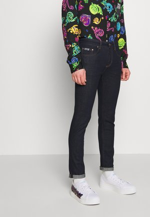 Homme portant un jean slim foncé, des baskets blanches à plateforme avec des détails colorés et une chemise noire à manches longues avec des motifs multicolores éclatants.