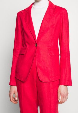 Femme portant un blazer rouge vif sur mesure avec un pantalon assorti et un col roulé blanc en dessous, debout devant un fond uni.