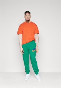 Ljusoranga bomullströja i kombination med gröna Nike sweatpants med en broderad logotyp. Modellen bär vita sneakers. Enkelt bakgrund.