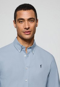 Light blue cotton button-up shirt with a collar, featuring a small navy golf figure logo on the left chest and button detailing.