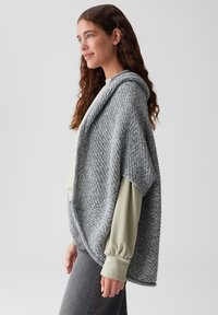 Grau gestrickter Poncho mit Kapuze, der über eine strukturierte Webart, einen lockeren Schnitt und weite Ärmel verfügt. Kombiniert mit einem hellgrünen Oberteil und dunklen Jeans.