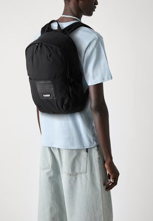 Zaino nero in materiale sintetico liscio, con doppie cerniere frontali, tasca laterale in rete e patch con logo, indossato sopra una maglietta azzurro chiaro.