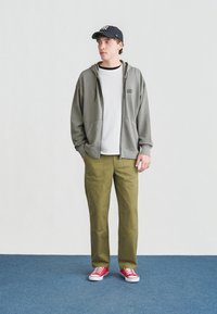 Νεαρός άνδρας με μαύρο ριγέ καπέλο, γκρι φούτερ, λευκό μπλουζάκι, χακί παντελόνι και κόκκινα αθλητικά παπούτσια, στέκεται σε μπλε χαλί μπροστά σε λευκό τοίχο.