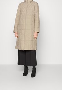 Abrigo acolchado beige con cuello alto y cremallera completa, con mangas largas y corte recto, llevado sobre pantalones negros de pierna ancha y botines hasta el tobillo.