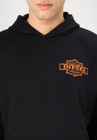 Čierna mikina s kapucňou vyrobená z bavlny, s predným klokanie vreckom. Obsahuje oranžovú nášivku loga Diesel na ľavej hrudi.