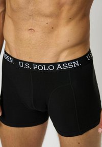 Črne bokserske hlače iz mehkega blaga z gladko teksturo. Elastični pas z logotipom prikazuje "U.S. POLO ASSN." v belem tisku.