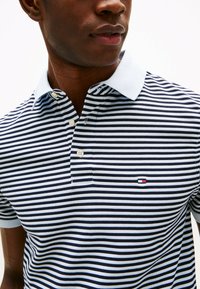 Stribet polo skjorte i navy og hvid, med en lyseblå krave, to knapper og et lille logo på venstre bryst.