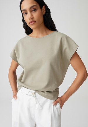 Femme aux longs cheveux noirs portant un haut beige ample à manches courtes et un pantalon blanc à cordon de serrage, debout les mains dans les poches.