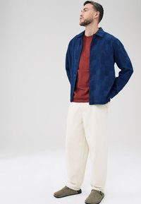 Camicia blu con motivo e colletto, t-shirt rossa a girocollo, pantaloni larghi color crema e scarpe slip-on grigie, in piedi davanti a uno sfondo neutro.