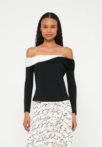 Õlavabad pika varrukaga mustast puuvillast pluus, millel on valge drapeeritud detail. Sujuv tekstuur, keha järgnev lõige ja kumeralt lõigatud serv.