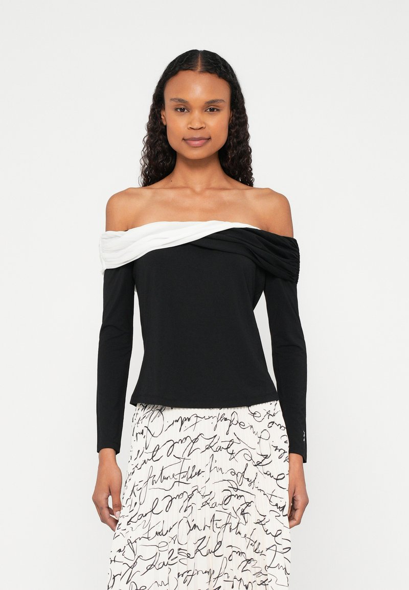 Õlavabad pika varrukaga mustast puuvillast pluus, millel on valge drapeeritud detail. Sujuv tekstuur, keha järgnev lõige ja kumeralt lõigatud serv.