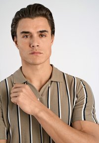 Beige randig polo-shirt med smala svarta och vita vertikala linjer, korta ärmar och en klassisk krage; modellen håller kragen med ena handen.