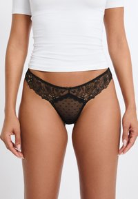Tanga de encaje negro con bordado floral, malla de topitos, bordes festoneados y sutiles acentos metálicos, mostrando una textura transparente.