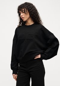 Μαύρο φούτερ με άνετη εφαρμογή, που διαθέτει υφή λογότυπου στο κέντρο, μανσέτες με ρίγες και στρογγυλή λαιμόκοψη. Συνδυάζεται με μαύρο παντελόνι.