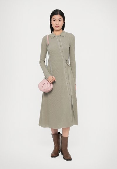 Robe longue côtelée vert olive avec fermeture à boutons, col et taille cintrée. Associée à un sac à main rose et des bottines marron.
