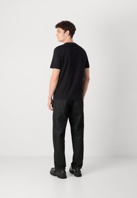 Zwarte t-shirt met korte mouwen en een ronde hals, gecombineerd met donkere, textuurrijke broeken en zwarte schoenen. Het model is van achteren bekeken.