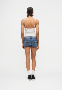 Άτομο με πλεξούδες που φοράει λευκό αμάνικο τοπ, σορτς τζιν, λευκές κάλτσες και μαύρα αθλητικά, στέκεται με την πλάτη σε απλό φόντο.